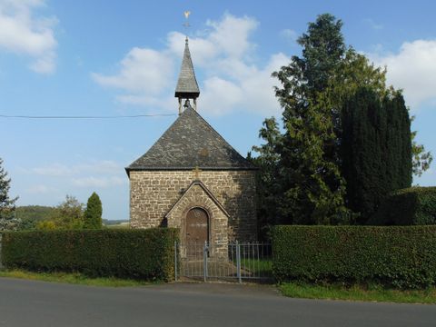 Eine kleine Steinkapelle mit spitzem Dach und Wetterhahn, umgeben von Bäumen und Hecken.