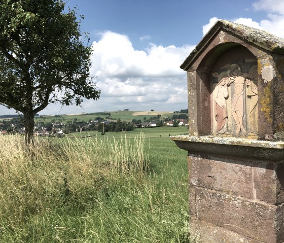 Ein Steinrelief am Wegesrand zeigt eine religiöse Szene. Im Hintergrund sind Felder, ein Baum und ein Dorf unter einem blauen Himmel zu sehen., © TI Bitburger Land