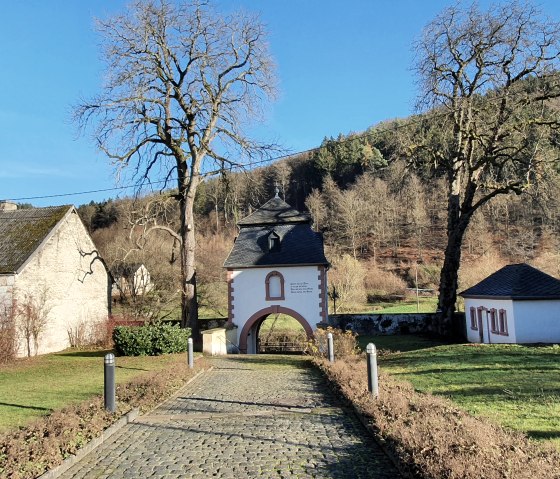 Entrance gate to the monastery complex of St. Thomas with paved path, surrounded by trees and buildings, in front of a wooded hill., © TI BItburger Land - Steffi Wagner