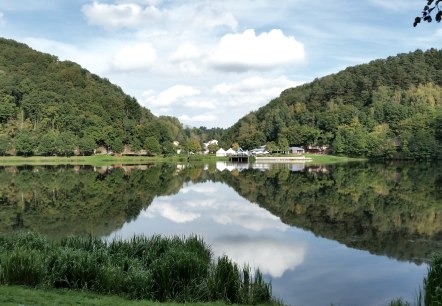 Panorama du lac de barrage de Bitburg avec des collines boisées, des bâtiments sur la rive et un reflet clair dans l'eau sous un ciel bleu., © TI Bitburger Land