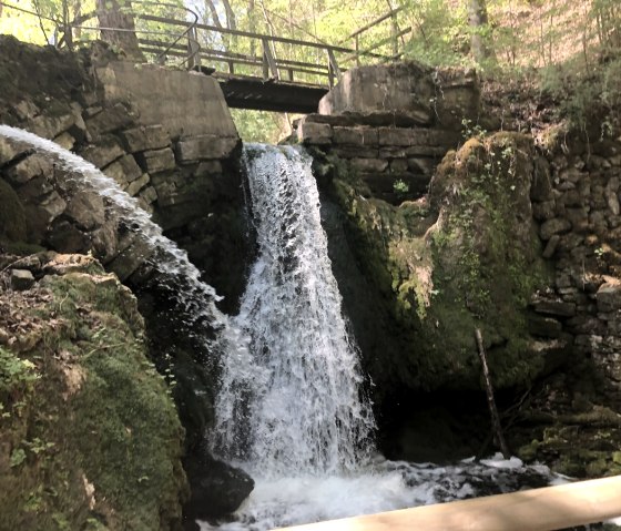 Ein Wasserfall fließt unter einer kleinen Brücke in einem bewaldeten Gebiet. Die Umgebung ist von Moos und Steinen geprägt., © TI Bitburger Land_Uschi Hallet