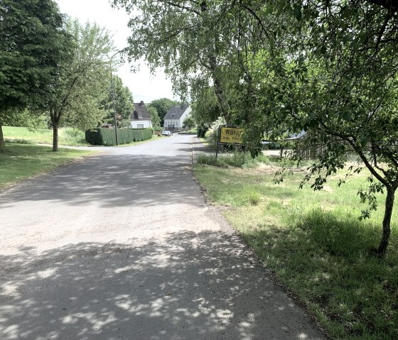 Straat in een dorp, omzoomd met bomen en een plaatsnaambord 'Röhl'. Huizen zijn te zien op de achtergrond., © Benjamin Milbach