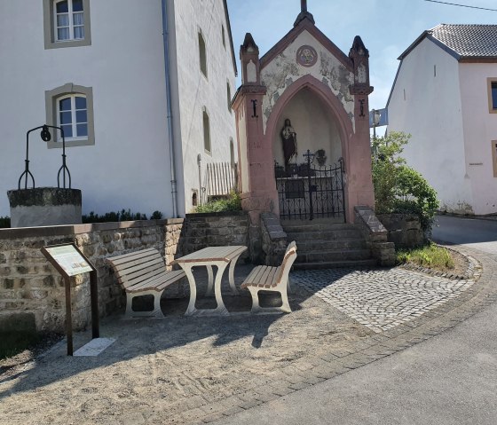 Kleine kapel met standbeeld en bank in Wolsfeld. Er is ook een fontein en een informatiebord. Zonnige dag., © Tourist-Information Bitburger Land, Steffi Wagner