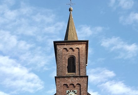 Backsteinkirche St. Laurentius mit Turm und Uhr, umgeben von Bäumen und einem Kreuz im Vordergrund, unter blauem Himmel., © TI Bitburger Land