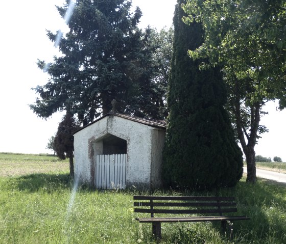 Kleine Kapelle neben einer Bank, umgeben von Bäumen und Wiese, an einem Feldweg., © TI Bitburger Land
