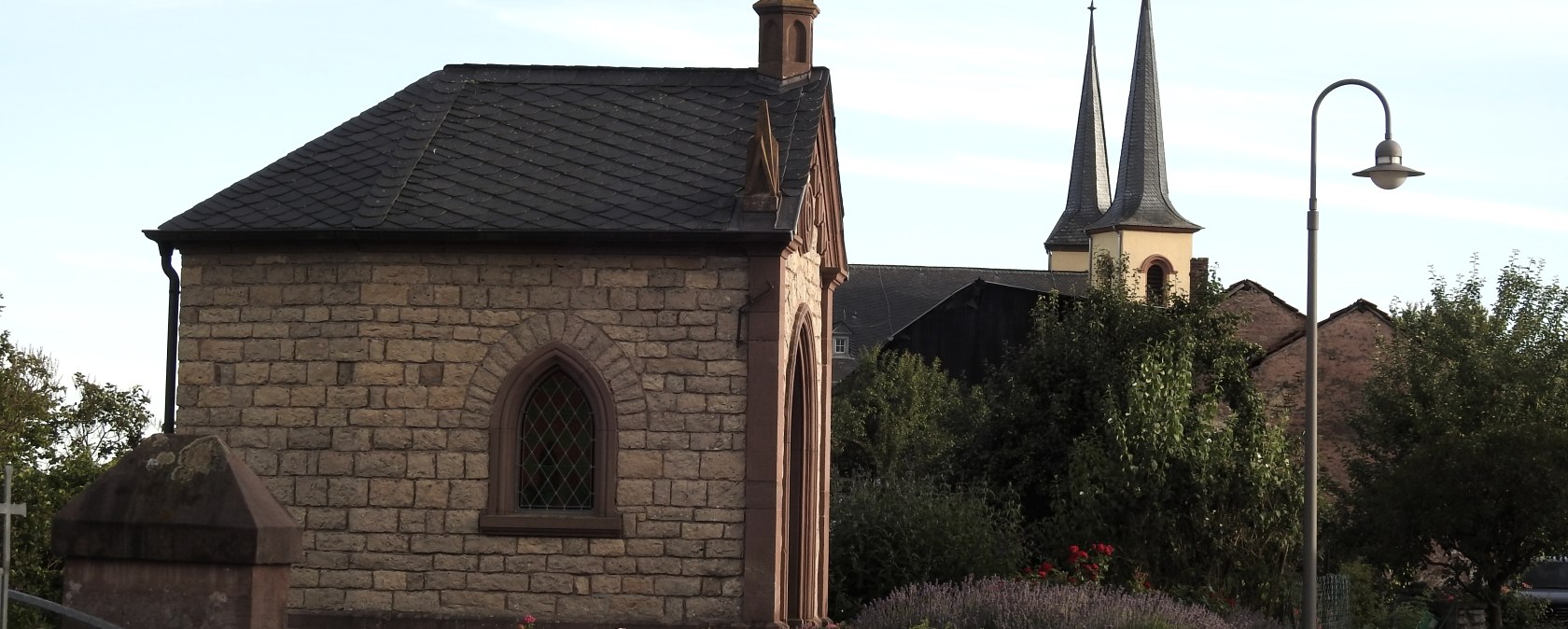 Chapelle en pierre avec un toit pointu et une fenêtre colorée, entourée de fleurs. En arrière-plan, on peut voir des clochers d'église., © Thomas Neises