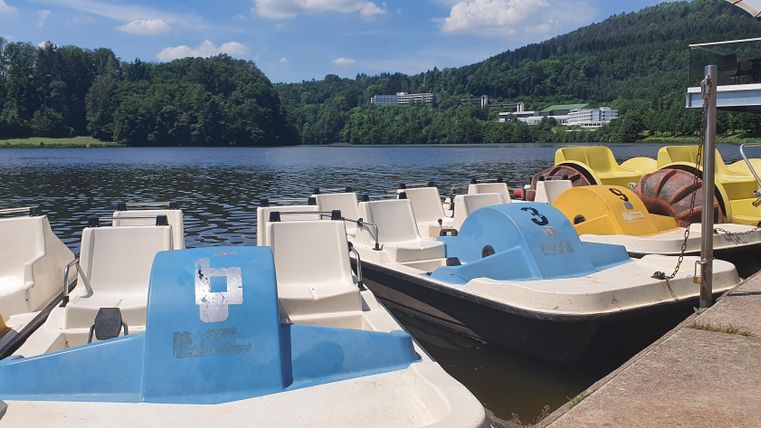 Ein ruhiger See mit mehreren Tretbooten am Ufer. Im Hintergrund sind grüne Berge und eine klare blaue Himmel zu sehen.