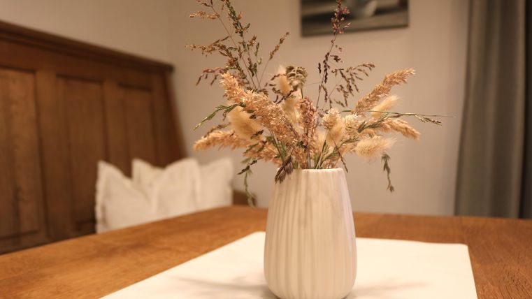 Een elegante vaas met gedroogde bloemen staat op een tafel.
Op de achtergrond zijn houten oppervlakten en een subtiele decoratie zichtbaar.