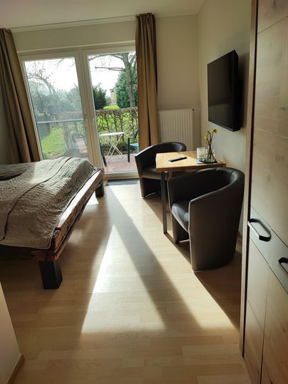 Ein helles Zimmer mit einem großen Bett und einem kleinen Tisch. Es gibt zwei Stühle und Fenster mit Blick auf einen Garten.