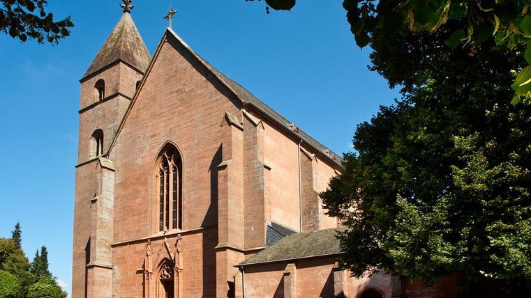 Eine beeindruckende Kirche aus rotem Stein unter einem klaren blauen Himmel. Umgeben von grünem Gras und Bäumen.