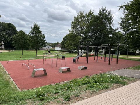 Un terrain de jeu avec des équipements de gymnastique sur un sol antidérapant. En arrière-plan, on peut voir des arbres verts et le ciel est nuageux.