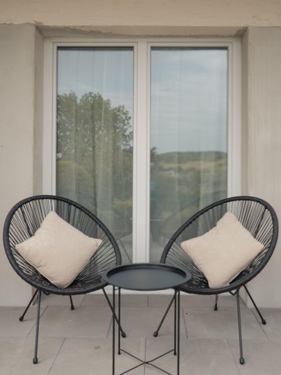 Ein gemütlicher Balkon mit zwei schwarzen Sesseln und einem kleinen Tisch. Die Sessel sind mit hellen Kissen ausgestattet, und im Hintergrund sind Fenster mit Vorhängen zu sehen.