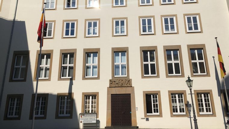 Ein historisches Gebäude mit mehreren Fenstern und zwei Fahnen. Die Fassade ist hell und klar, umgeben von einem Platz.