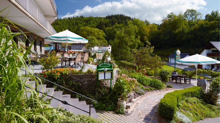 Terrasse des Hotel Haus Kylltal mit Tischen und Sonnenschirmen, umgeben von grüner Landschaft.