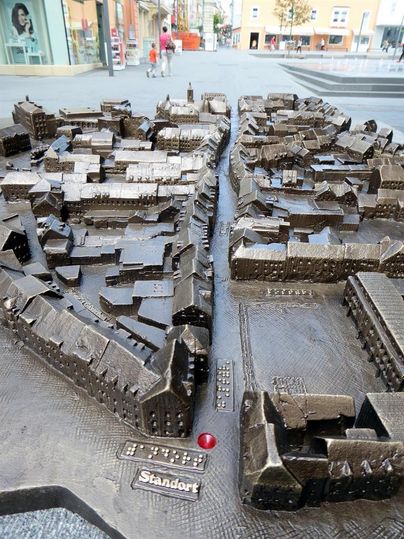 Eine detaillierte Stadtmodell aus Metall zeigt die Anordnung von Gebäuden und Straßen. Es ist auf einem Platz aufgestellt und bietet eine Übersicht über die Umgebung.