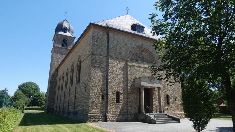 A historic church with a stone facade and a bell tower. The sky is clear and the surroundings are green and sunny.