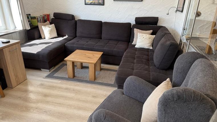 Ein gemütliches Wohnzimmer mit einem großen, bequemen Sofa und einem kleinen Holztisch. An der Wand hängen einige Bilder und es gibt viel Tageslicht.