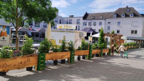 Une place animée avec un banc en bois et des plantes. À l'arrière-plan, on peut voir des bâtiments et des étals.