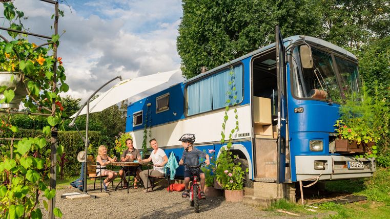 Ein blauer Bus steht in einer grünen Umgebung. Eine Familie sitzt am Tisch im Freien und genießt die Zeit miteinander.