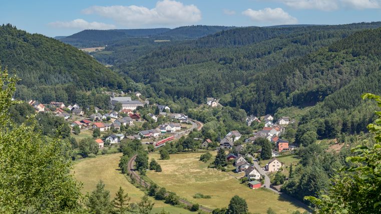 Blick auf ein Dorf in einem bewaldeten Tal mit Hügeln im Hintergrund.