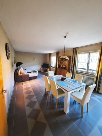 Ein helles Esszimmer mit einem großen Tisch und weißen Stühlen. Im Hintergrund befindet sich ein gemütlicher Wohnbereich mit einem Sofa und einem Fernseher.