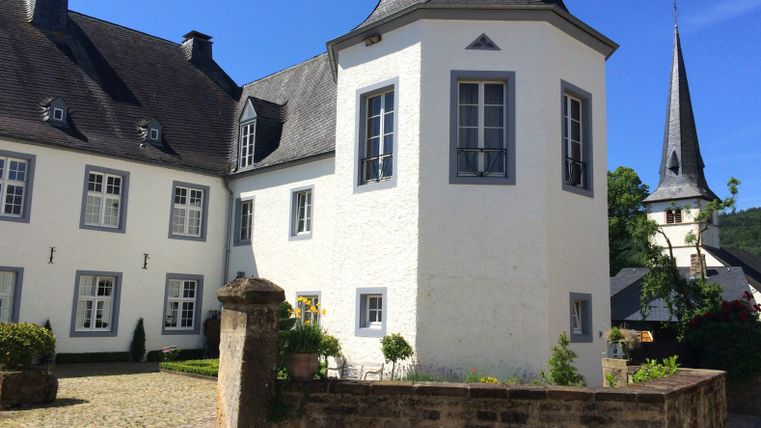 Weißes Schlossgebäude mit grauem Dach und Turm, umgeben von Garten und Steinmauer, bei blauem Himmel.