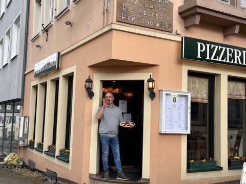 Ein Mann steht vor einem Pizzarestaurant und hält ein Tablett mit Speisen in der Hand. Das Gebäude ist freundlich gestaltet und hat große Fenster.