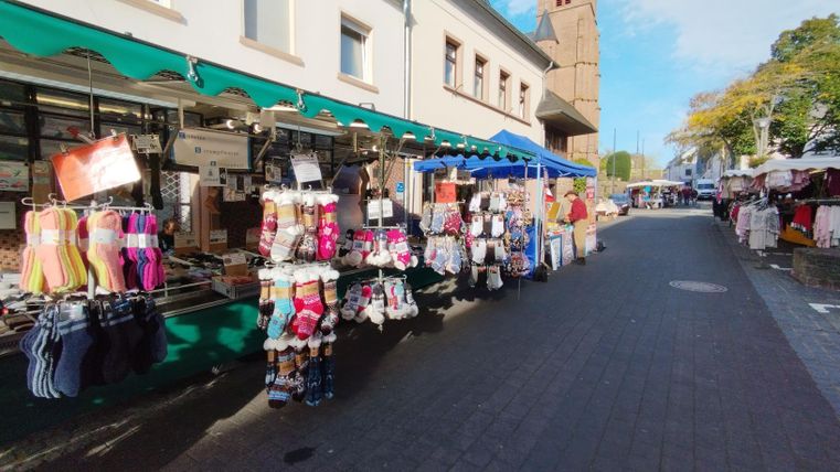 Een markt met kleurrijke kledingstands en tenten. De straat is druk en omgeven door gebouwen.