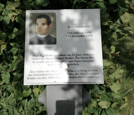 Plaque commémorative pour Peter Friedhofen avec photo et texte, entourée de feuilles vertes., © TI Bitburger Land