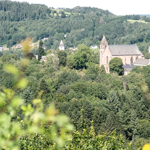 Gezicht op een kerk, omringd door dichte bossen en heuvels, onder een blauwe lucht. Op de voorgrond zijn vage planten te zien., &copy; Tourist-Info Bitburger Land M.Mayer