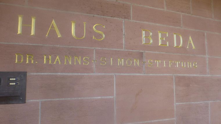 Eine Wand mit goldenem Schriftzug. Es steht "HAUS BEDA" und "DR. HANNS SIMON STIFTUNG".