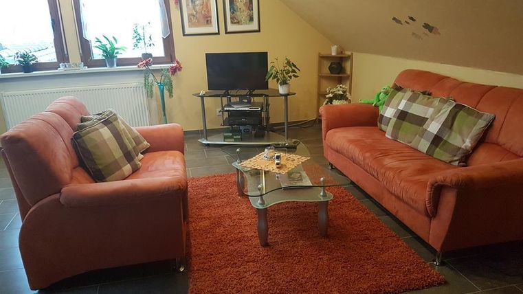 Ein gemütliches Wohnzimmer mit zwei orangenen Sofas und einem niedrigen Glastisch. Im Hintergrund stehen Pflanzen und ein Fernseher auf einem Regal.