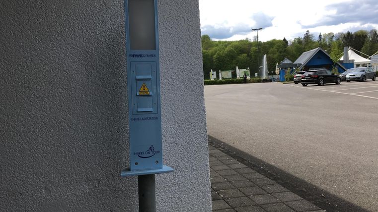 Un parcmètre bleu est adossé à un mur. En arrière-plan, un parking avec des véhicules et la nature environnante sont visibles.