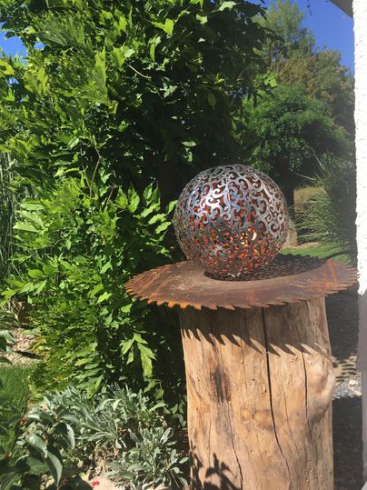 Een decoratieve metalen bol staat op een houten stam, omringd door groene planten. De achtergrond toont een zonnig tuinenlandschap.