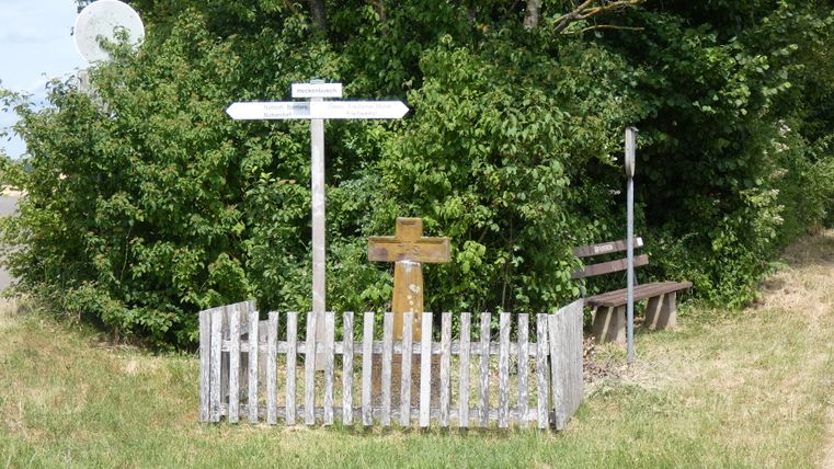 Wegweiser und Holzkreuz auf einem Wanderweg, umgeben von Büschen.
