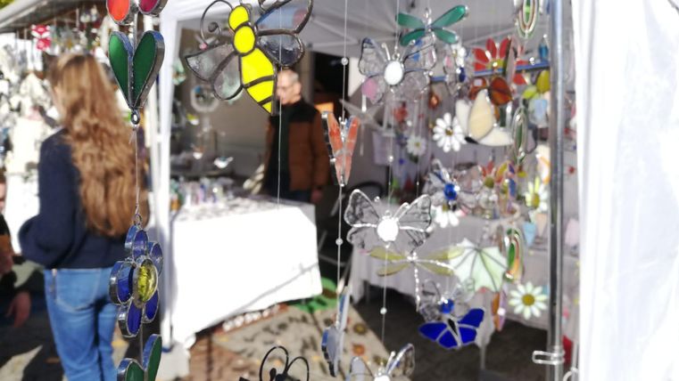 Colorful glass decorations hang at a stand in a market. In the background, people and more goods can be seen.