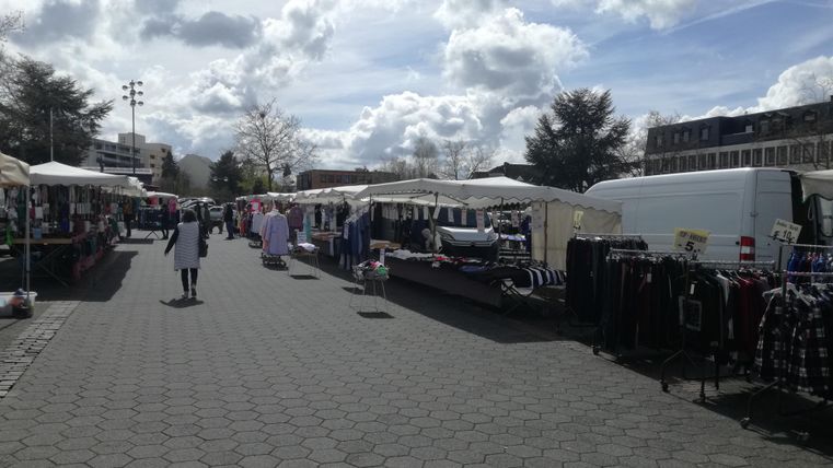 A bustling market with stalls and numerous goods. The sky is cloudy, and many people are strolling around.