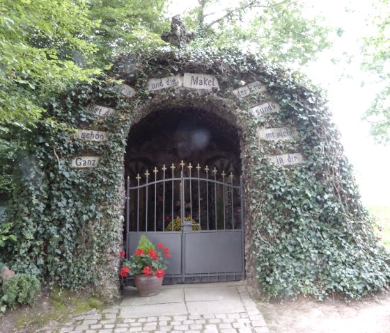 Une grotte de Lourdes recouverte de lierre avec une porte. Des fleurs sont visibles sur le portail. Des panneaux avec des inscriptions décorent l'entrée., © Eifelverein Ortsgruppe Speicher