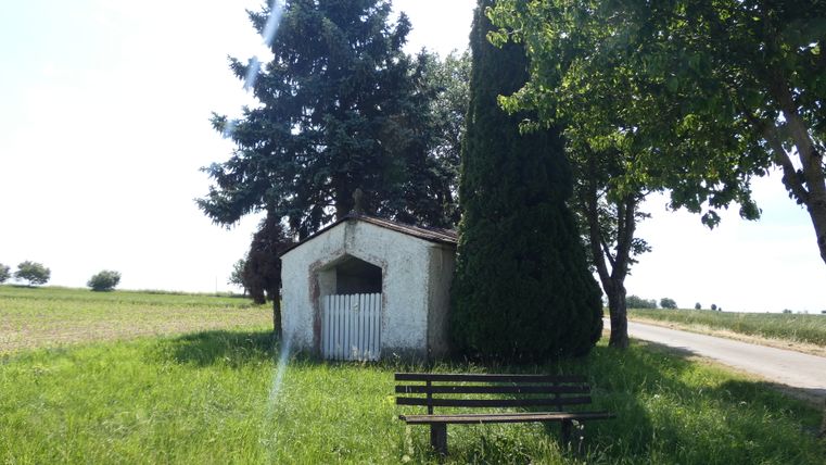 Eine kleine Kapelle neben einer Bank und Bäumen auf einem Feldweg.