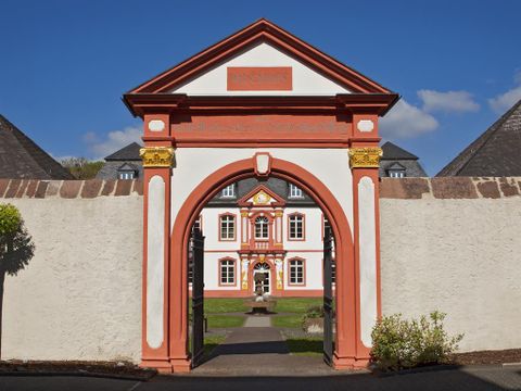 Ein historisches Gebäude mit roten und weißen Wänden ist durch einen großen Bogen sichtbar. Die Umgebung ist grün und gepflegt, unter einem klaren blauen Himmel.