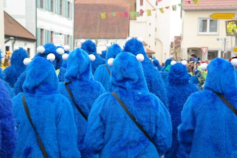 Eine Gruppe von Menschen in blauen Kostümen mit großen Augen, die an ein lustiges Monster erinnern. Sie stehen bei einer festlichen Parade in einer Stadt.