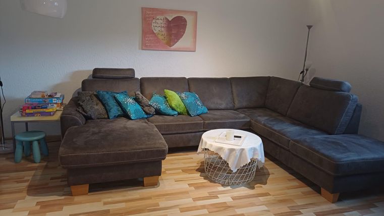 A cozy living room with a large gray sofa and colorful cushions. Next to it is a table with a tablecloth and books on a small table.
