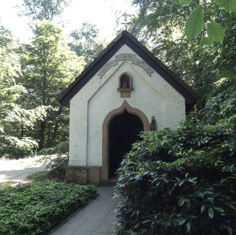 Eine kleine Kapelle aus dem Jahr 1877 steht im Wald, umgeben von Bäumen und einem Weg. Die Sonne scheint durch das Blätterdach.