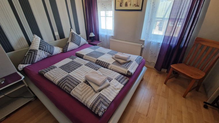 Ein gemütliches Schlafzimmer mit einem Doppelbett und gestreiften Wänden. Helle Vorhänge lassen viel Licht herein und ein Holztischstuhl steht neben dem Bett.