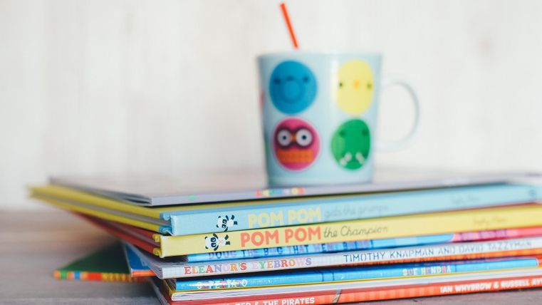A stack of colorful children's books is lying on a table. On top of it is a cup with cheerful faces and a straw.
