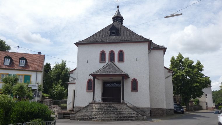 Kirche in Fließem mit weißer Fassade und rotem Dach, umgeben von Bäumen und Häusern.