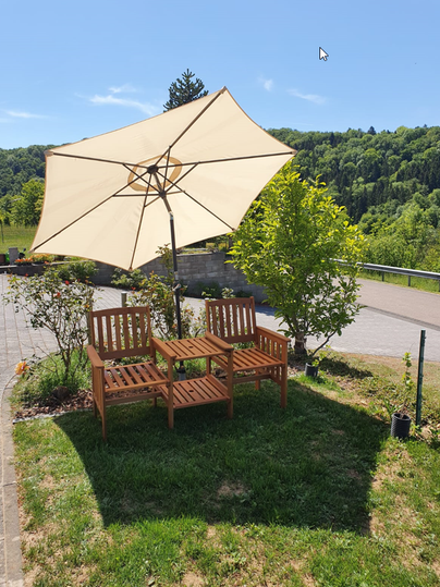 Ein gemütlicher Sitzbereich mit Holzmöbeln und einem großen Sonnenschirm. Umgeben von grünen Pflanzen und einem klaren blauen Himmel.
