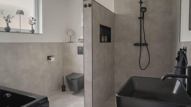 Ein modernes Badezimmer mit grauen Fliesen und eleganten schwarzen Sanitäranlagen. Die Dusche ist offen und gut beleuchtet, mit einem Fenster, das Tageslicht hereinlässt.