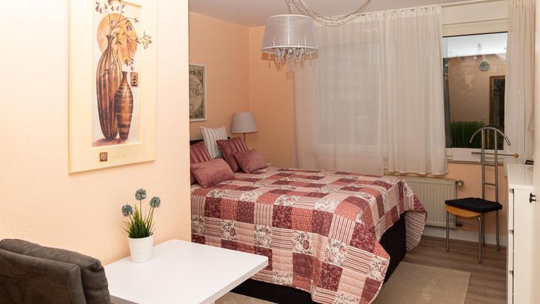 Ein gemütliches Schlafzimmer mit einem Doppelbett und einer hellen, warmen Farbgestaltung. Es gibt einen kleinen Tisch und einen Stuhl, sowie Fenster mit Vorhängen.