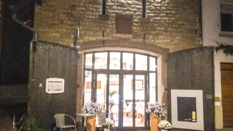 Ein schön beleuchtetes Gebäude mit großen Fenstern und einer einladenden Eingangstür. Vor dem Gebäude stehen Tische und Stühle, um einen gemütlichen Außenbereich zu schaffen.